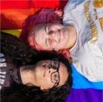 Two people - one with pink hair, the other with long black hair and glasses - lay on a rainbow pride flag and grin up at the camera.